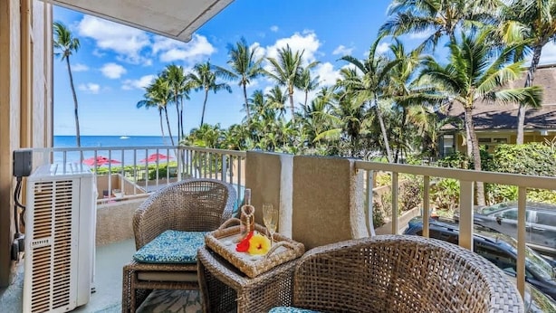 A balcony with wicker furniture, a small table with a drink, and a view of the ocean and palm trees. - 1 bedroom Beachfront Condo, AC, Lanai Oceanview, 6 mile sandy beach (Kihei)