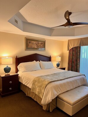 A hotel room with a large bed, two bedside tables with lamps, a ceiling fan, a framed picture on the wall, and a window with curtains.