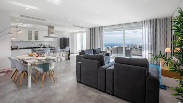A modern living room with a grey sofa, a dining area with a white table and chairs, and large windows offering a view of the city. - Casa Chloe - Luxury Ocean View Apartment (Lagos)