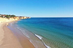 Ein Strand mit klarem, blauem Wasser, sandigem Ufer und einer Klippe mit Vegetation.
