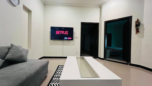 Ein modernes Wohnzimmer mit einem grauen Sofa, einem an der Wand befestigten Flachbildfernseher, der 'NETFLIX' zeigt, einem weißen Couchtisch und einem gemusterten Teppich.
