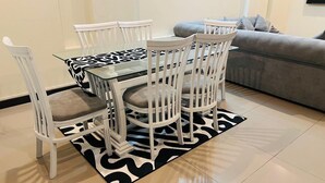 Ein Essbereich mit einem Tisch aus Glasplatte und weißen Stühlen, einem gemusterten Teppich und einem grauen Sofa im Hintergrund.