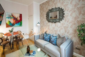 A living room with a sofa, coffee table, and chairs. There is a painting on the wall and a mirror above the sofa.