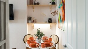 A dining area with a round table set for four, surrounded by wicker chairs. Above the table, there are two wooden shelves with decorative items and a painting. - Charming 1-bed Apartment in London (London)