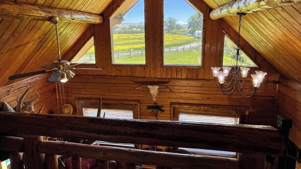 A log cabin with a sloped roof, wooden walls, a ceiling fan, a chandelier, and a large window offering a view of a green landscape. - The Caddis House- Big Hole River (Melrose)