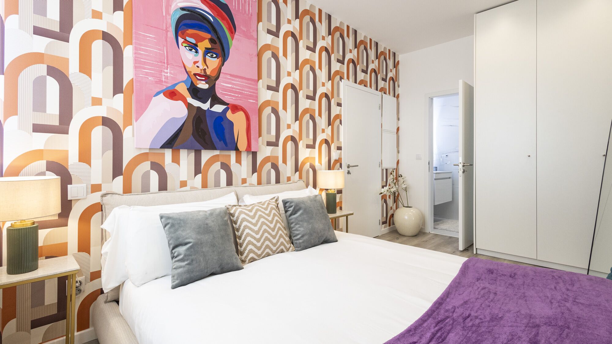 A modern bedroom with a large bed, a colorful abstract painting, and a geometric patterned wall.