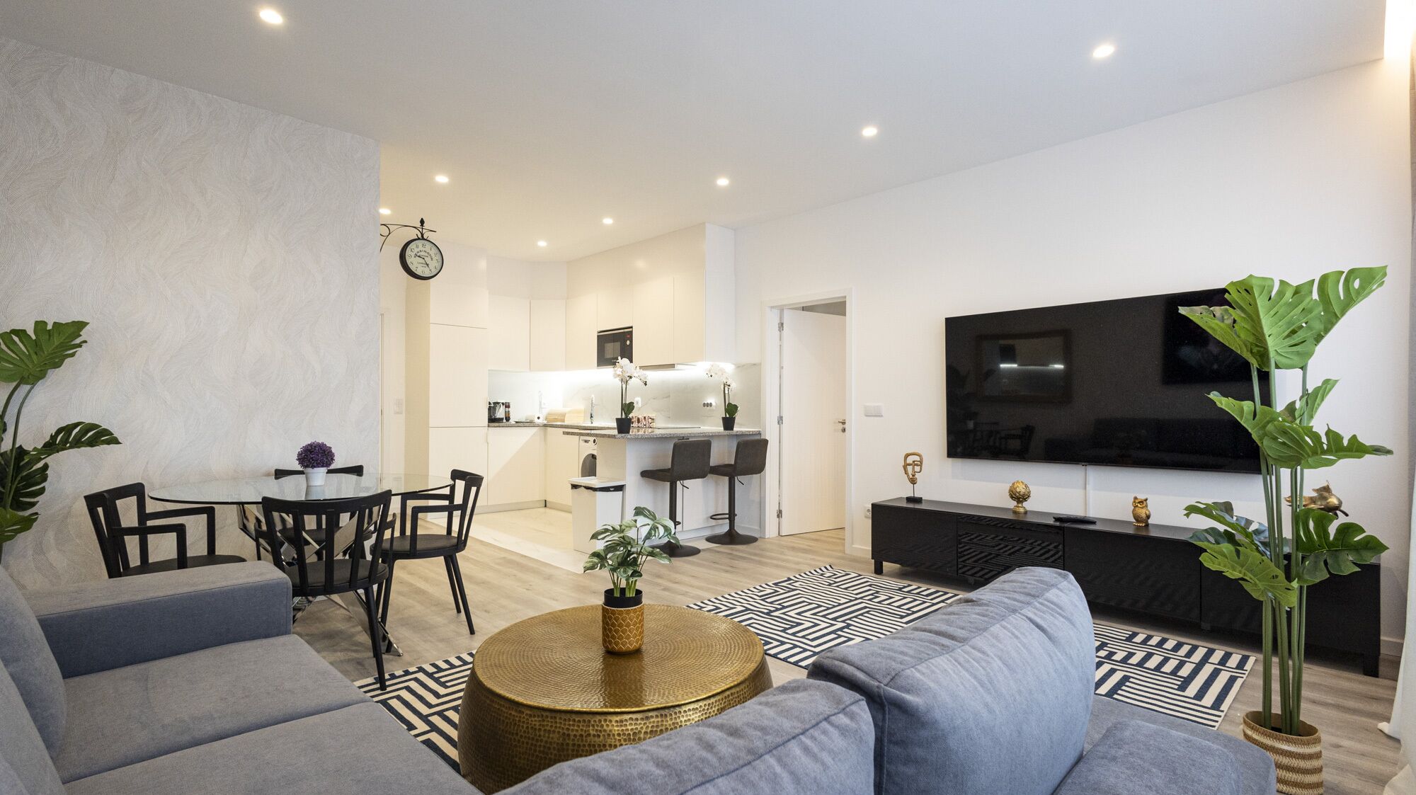 A modern living room with a sofa, coffee table, and TV. There is a dining area with chairs and a table. The kitchen is visible in the background.