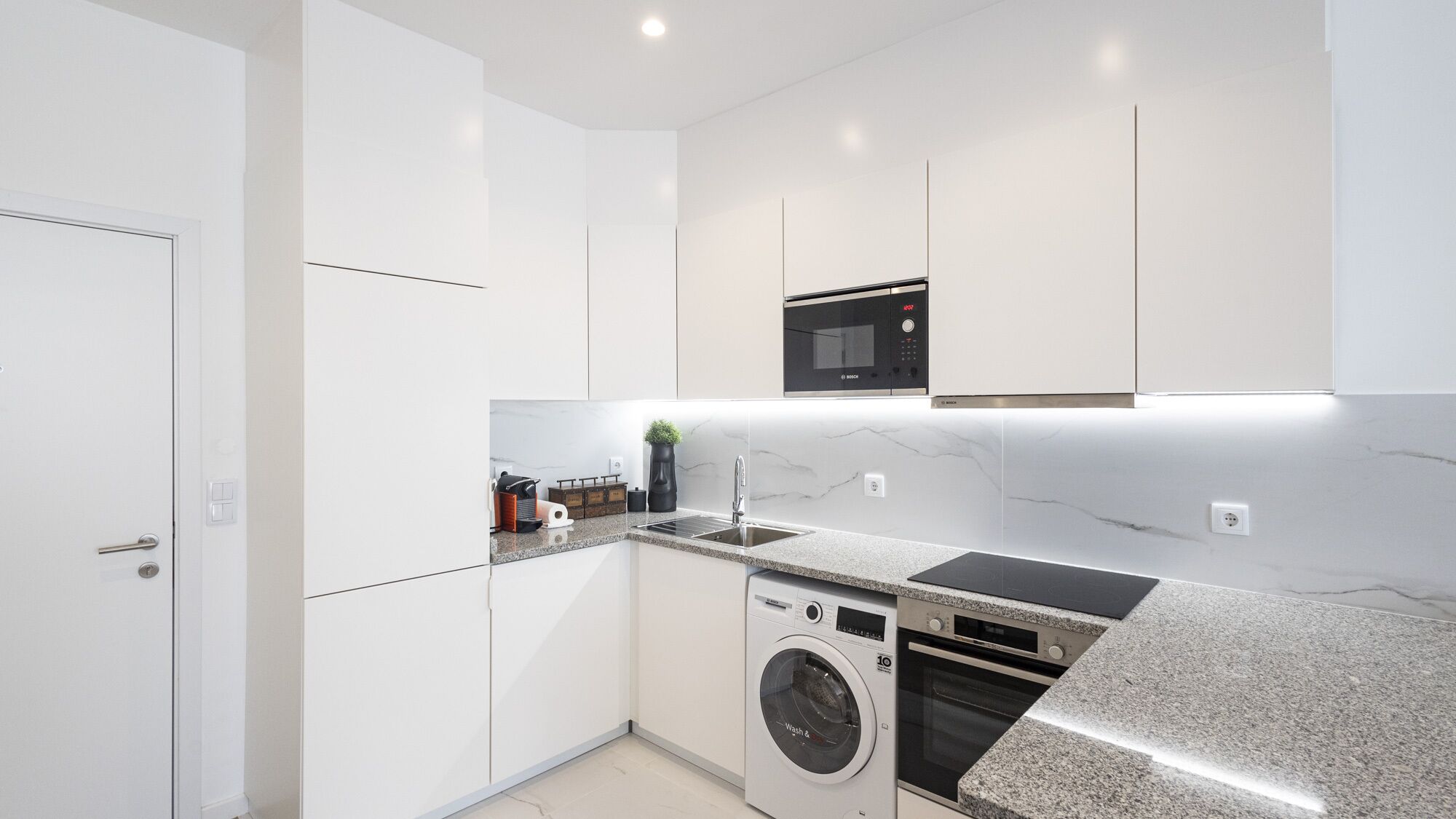 Una cocina moderna con armarios blancos, horno empotrado, microondas y lavadora.