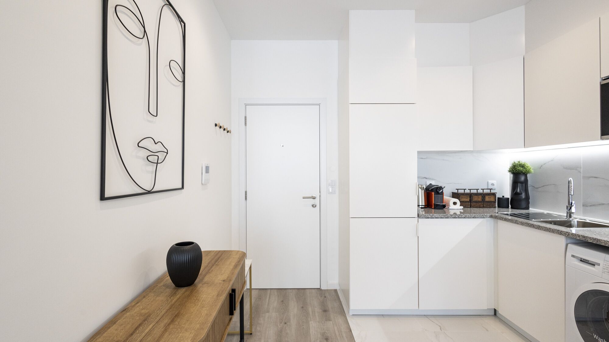 A modern kitchen with white cabinets, a wooden table with a black vase, and a wall-mounted abstract art piece.