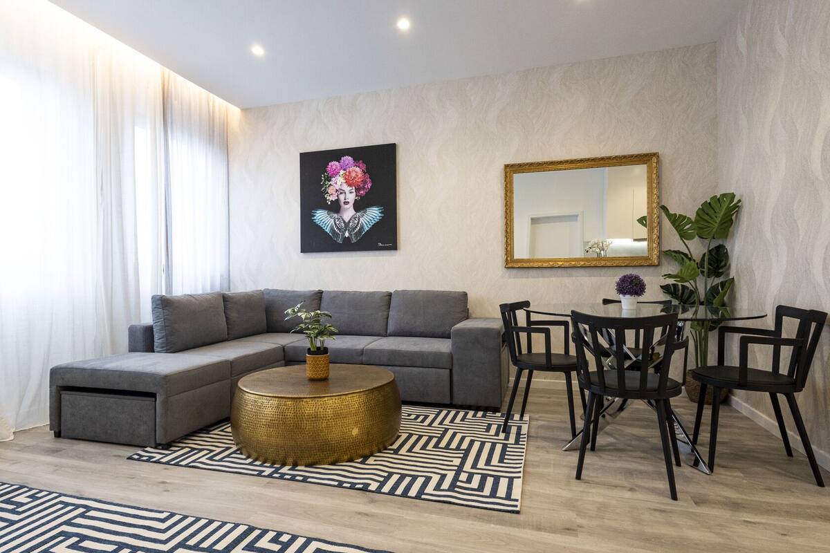 A modern living room with a grey sofa, a round gold coffee table, and a dining area with black chairs and a small table.
