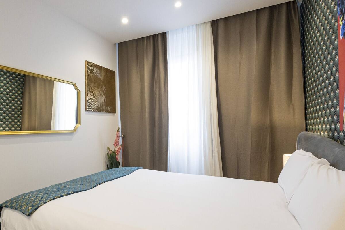 A hotel room with a bed, a large mirror, a window with curtains, and a wall-mounted artwork.