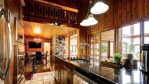 A wooden cabin interior with a stone fireplace, a kitchen with granite countertops, and a dining area with a stone wall. - Authentic Swiss Cabin - 4 BR with Hot Tub and pool table (Sainte-Agathe-des-Monts)