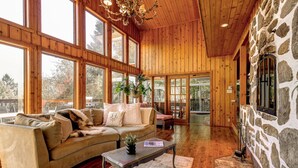 A spacious living room with a wooden ceiling, large windows, a stone wall, and a cozy sofa. - Authentic Swiss Cabin - 4 BR with Hot Tub and pool table (Sainte-Agathe-des-Monts)
