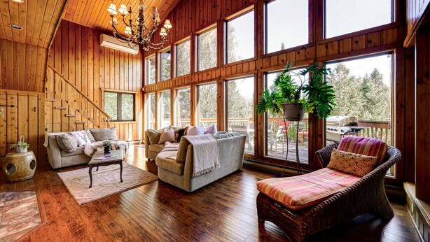 A spacious room with wooden walls and large windows, furnished with a sofa, armchairs, and a wicker chair. - Authentic Swiss Cabin - 4 BR with Hot Tub and pool table (Sainte-Agathe-des-Monts)
