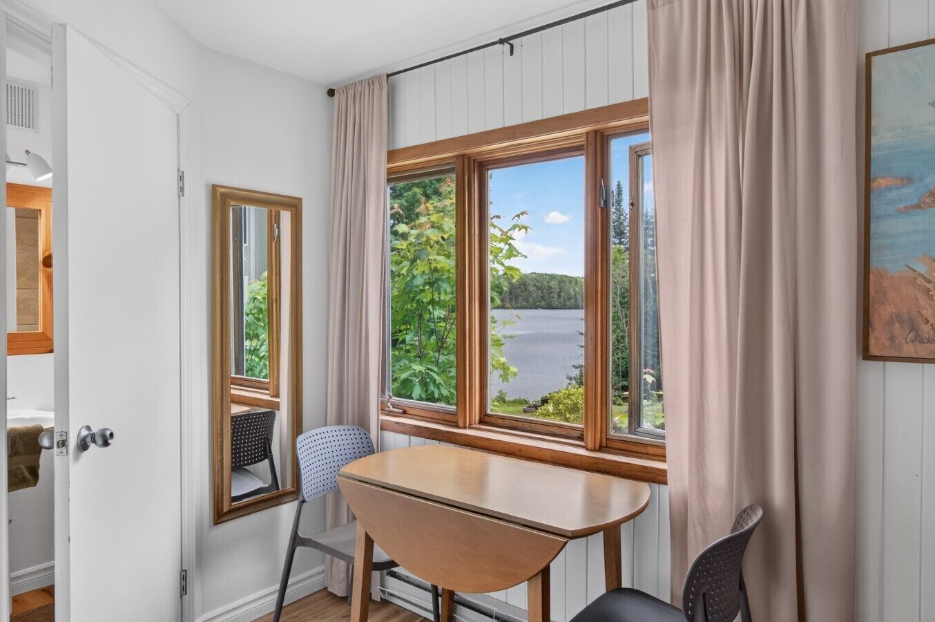 A room with a wooden table, two chairs, a window with a view of a lake and trees, and a door with a mirror.