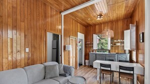 A modern interior with wooden walls, a grey sofa, a dining area with a table and chairs, and a kitchen with a window view. - Chalet Boréal, SPA, LAC & SKI (Saint-Donat-de-Montcalm)