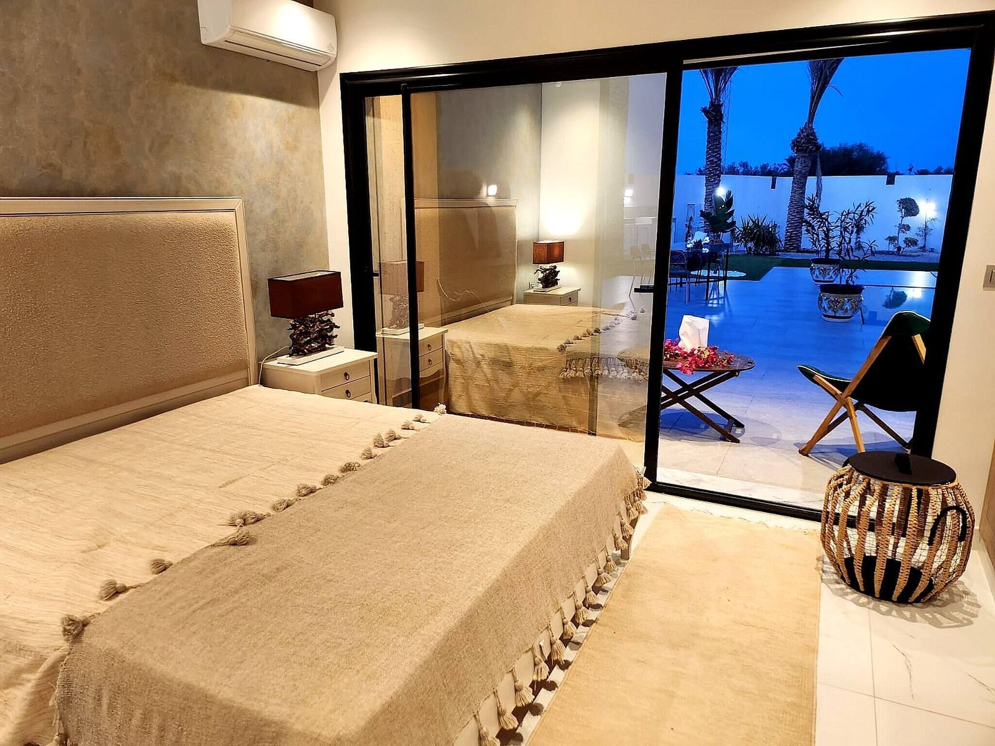 Un dormitorio con una cama grande, una mesita de noche, una lámpara y vistas a un patio exterior con sillas y una mesa.