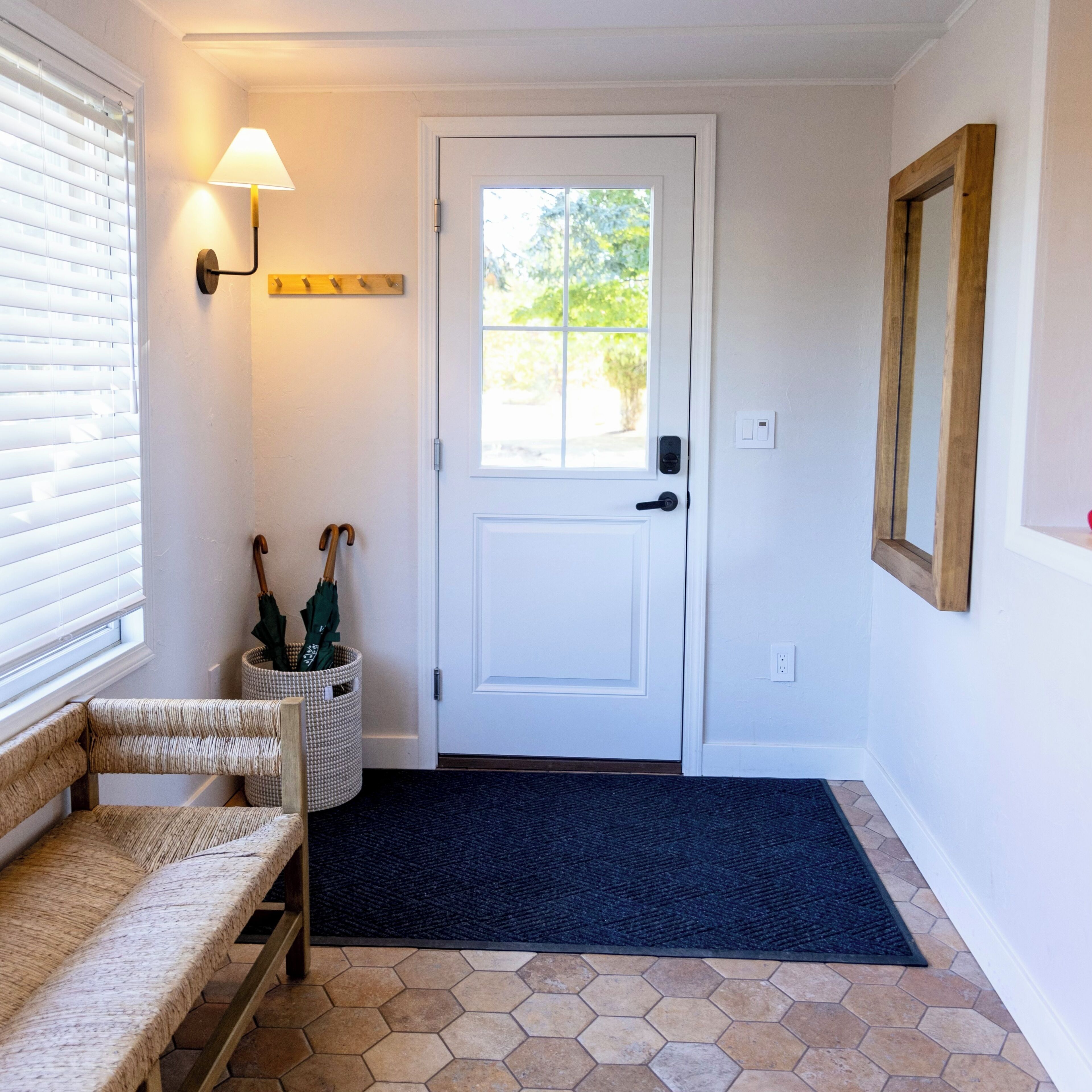Un pasillo con una puerta blanca, un banco, una lámpara de pared y una ventana con persianas.