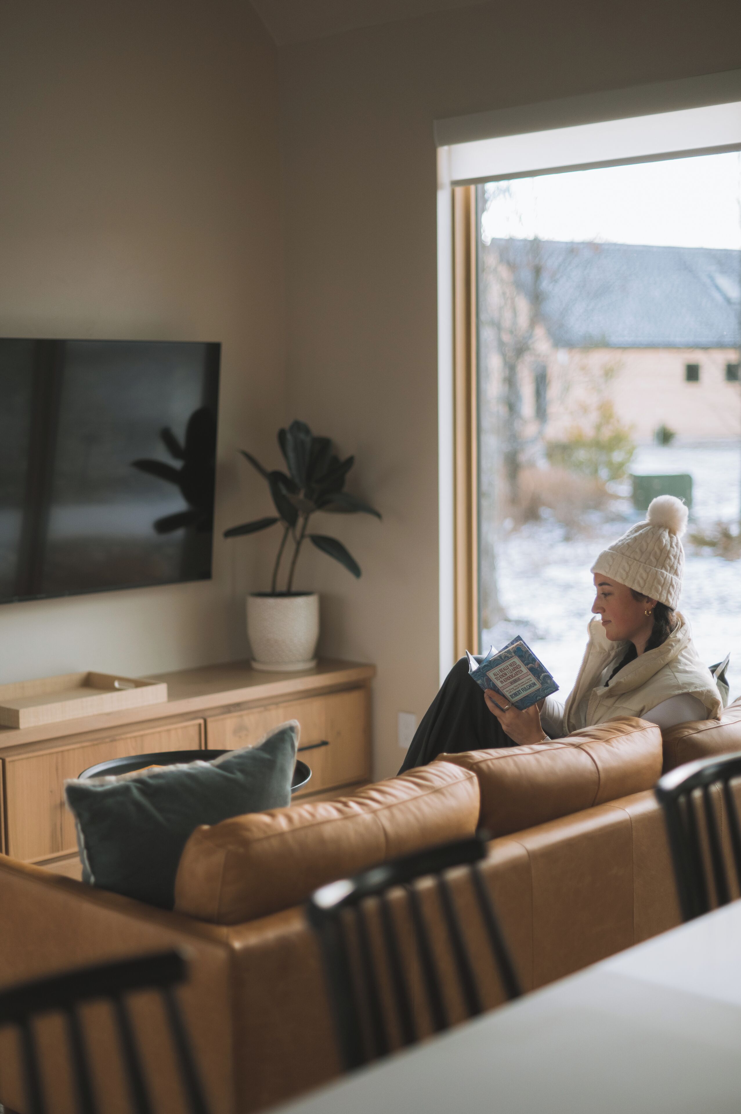 A person sitting on a leather couch reading a book in a room with a large window, a potted plant, and a flat-screen TV mounted on the wall.