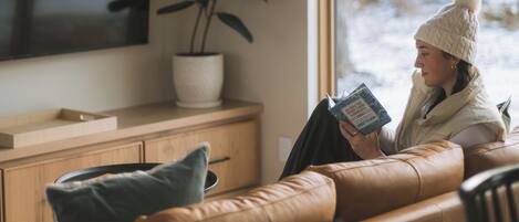 A person sitting on a leather couch reading a book in a room with a large window, a potted plant, and a flat-screen TV mounted on the wall.