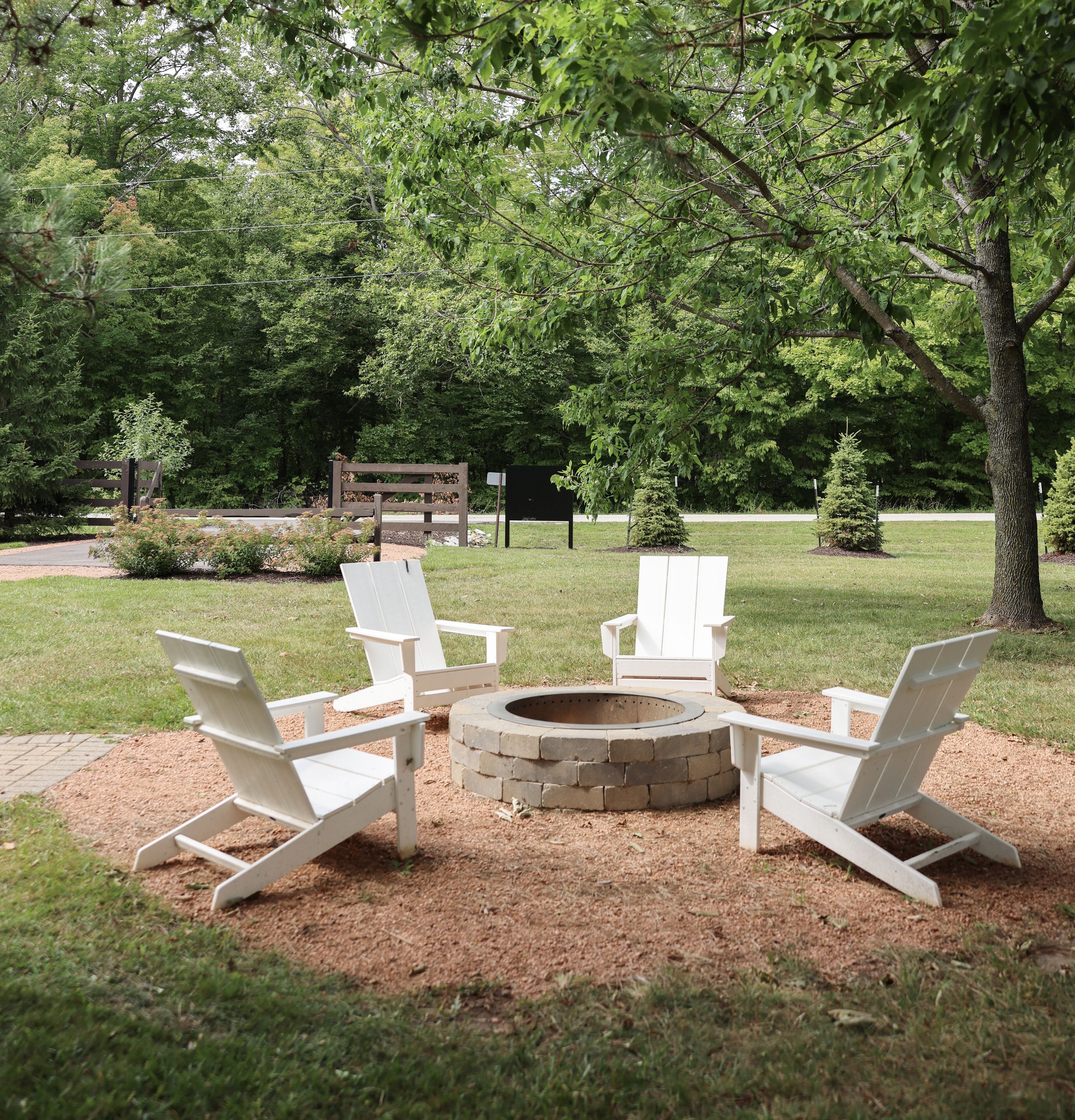 Una fogata rodeada de cuatro sillas Adirondack en un jardín ajardinado con árboles y un banco de madera al fondo.