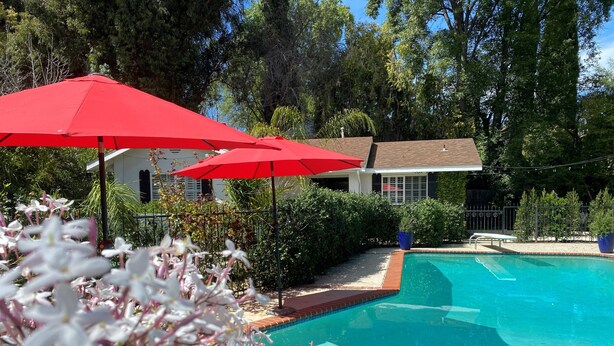 Piscina com um guarda-sol vermelho, rodeada por vegetação e uma casa ao fundo.