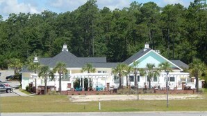 A two-story building with a light blue roof, surrounded by palm trees and a forested area. - Beautiful home in Garden City SC golf course community with easy access to beach (Murrells Inlet)