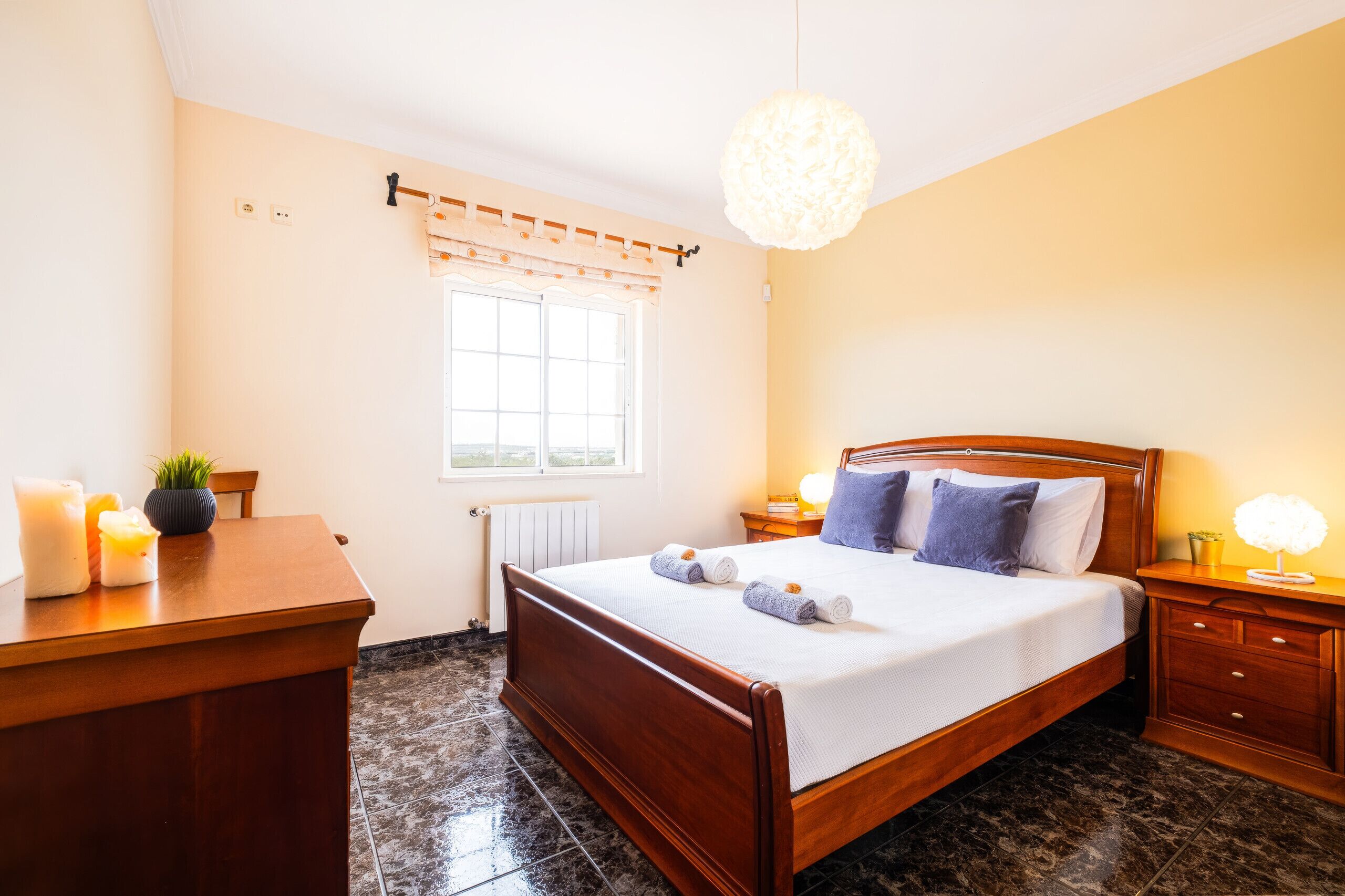 A bedroom with a wooden bed, bedside tables, a window with curtains, a radiator, and a hanging light fixture.