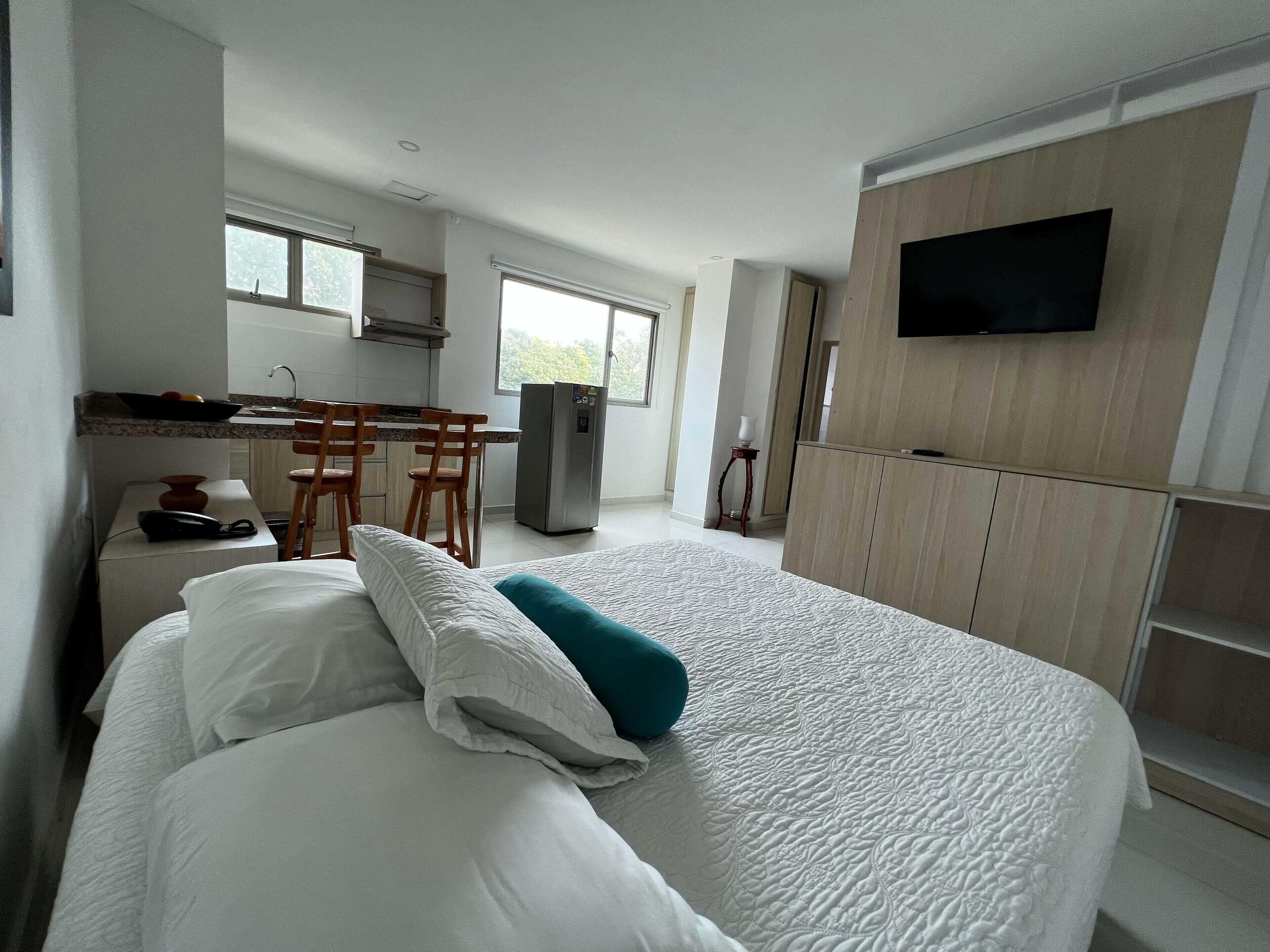 A hotel room with a large bed, a desk with chairs, a refrigerator, a television mounted on the wall, and a window providing natural light.