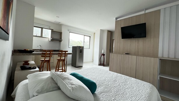 A modern hotel room with a bed, a TV mounted on the wall, a kitchenette, and a large window.