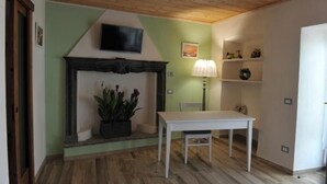 Una habitación con chimenea, un televisor montado encima, una mesa con una silla y una lámpara sobre una mesita.