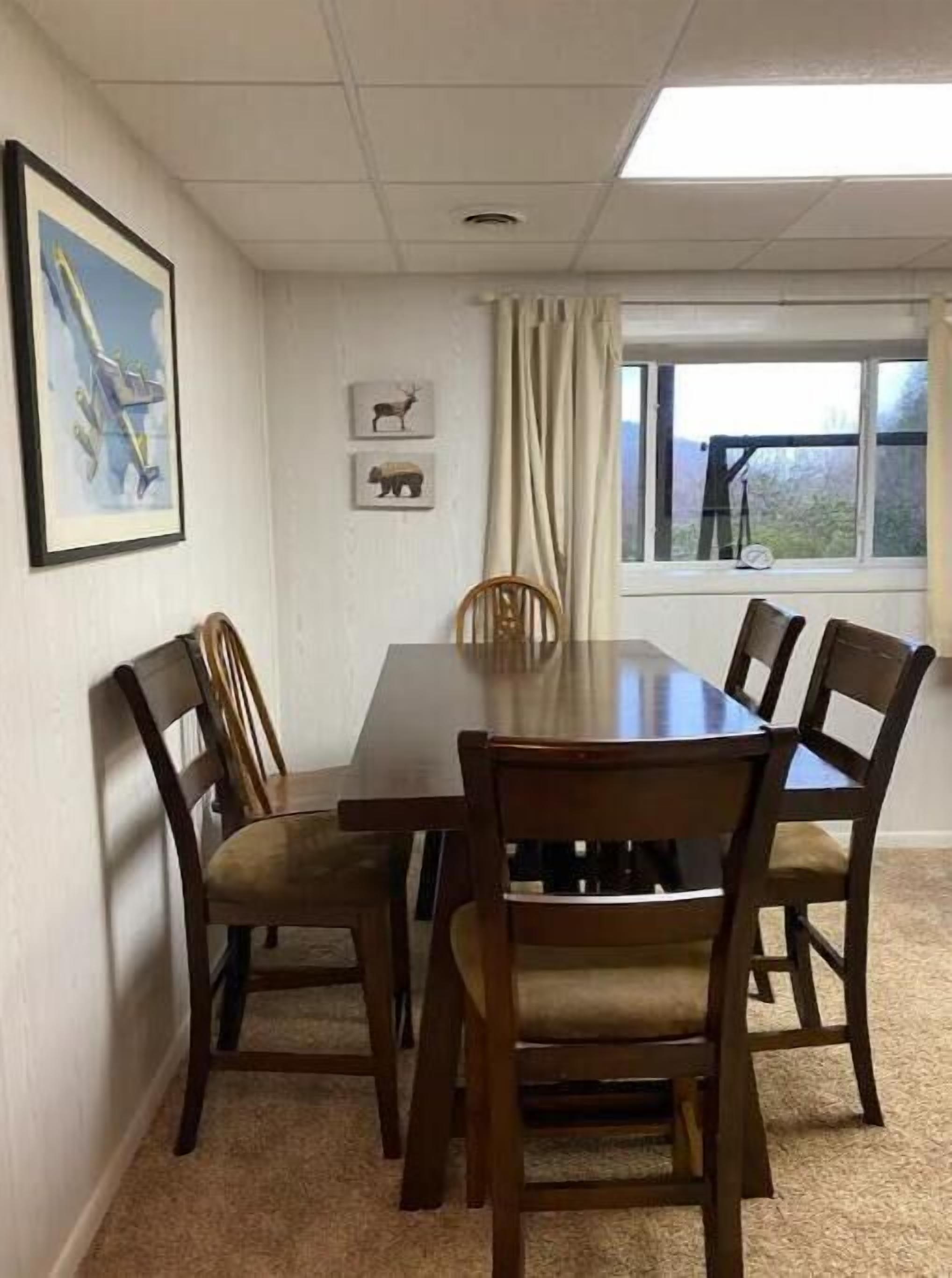 Comedor con mesa de madera y cuatro sillas, una ventana con cortinas y dos cuadros en la pared.