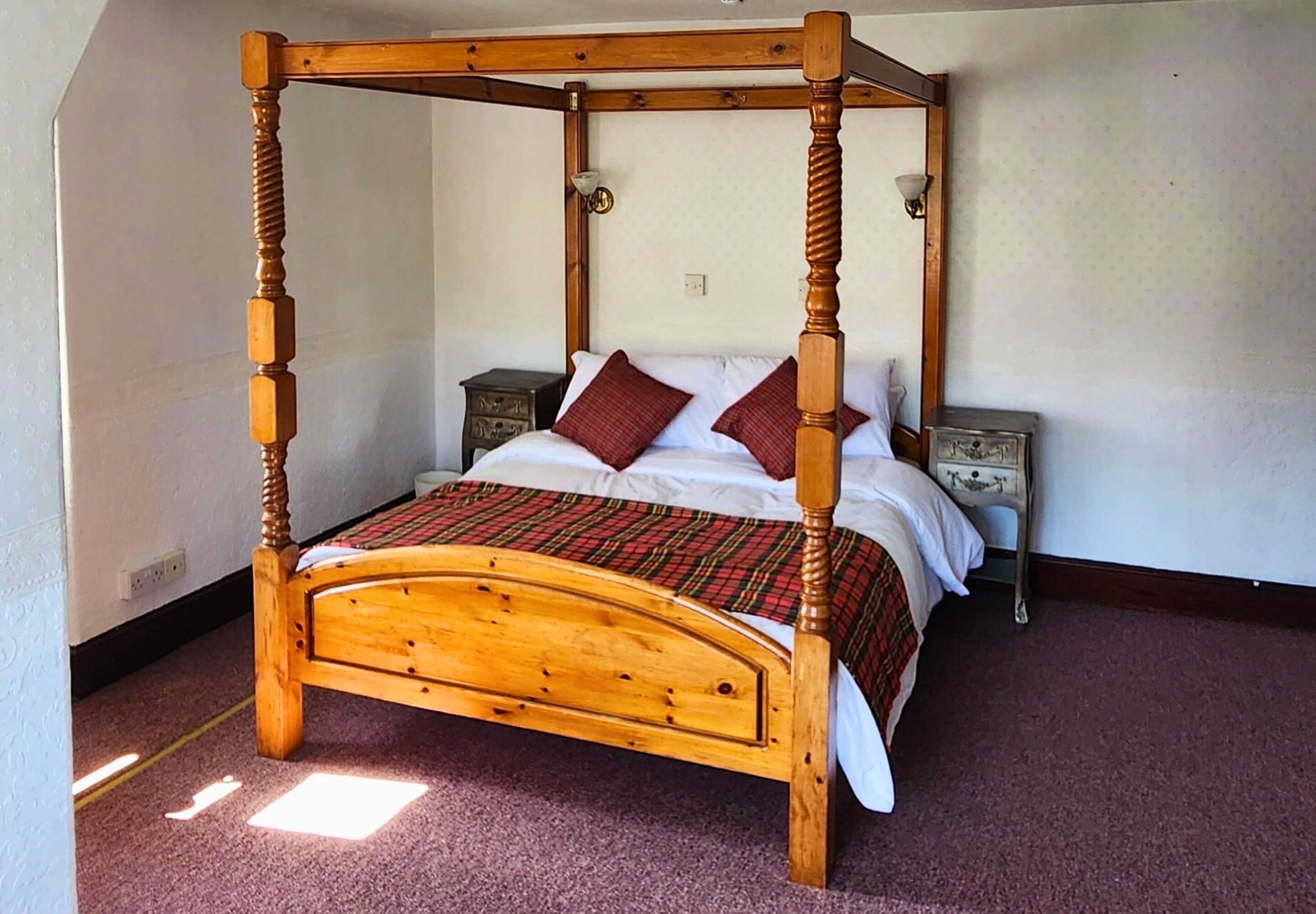 A four-poster bed with a plaid bedspread and two pillows, a nightstand with a lamp, and a wall socket.