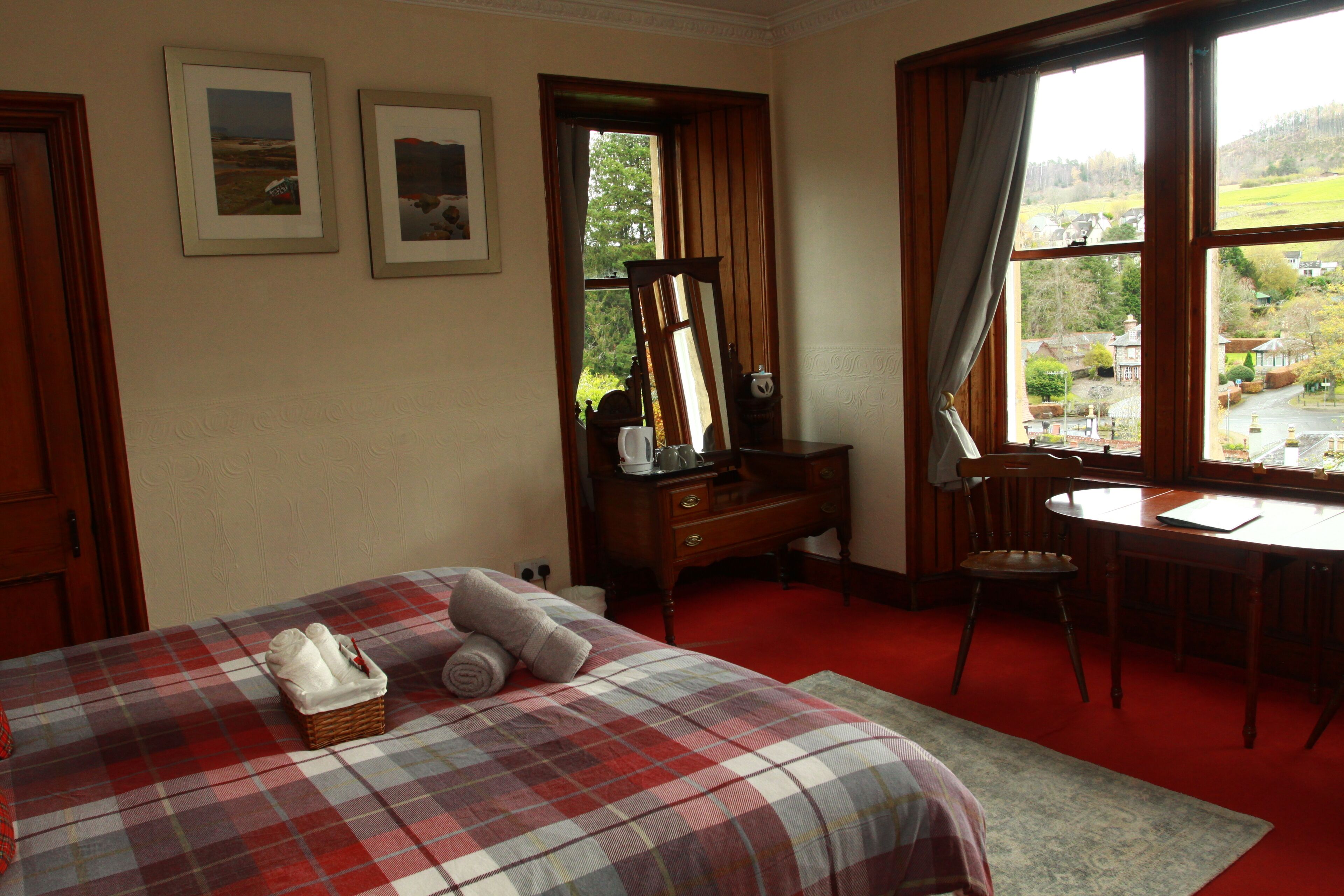 A hotel room with a bed, a desk, a chair, a window with a view, and a red carpet.