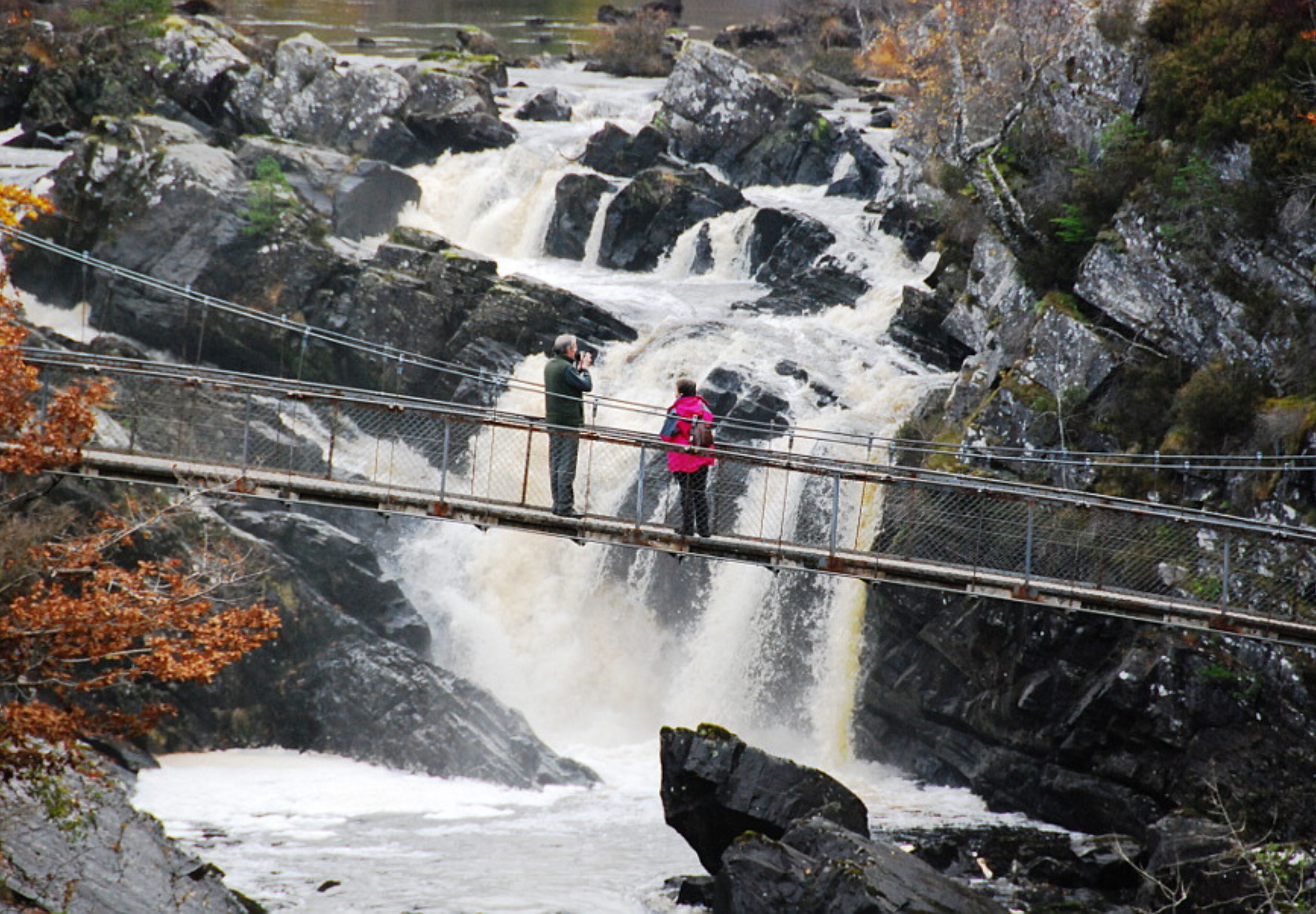 A suspension bridge over a waterfall with rocky surroundings and a person in a pink jacket.