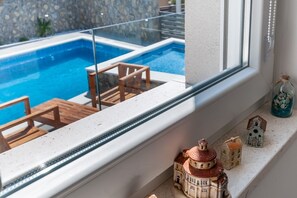 Une vue par la fenêtre sur une piscine, séparée par une barrière en verre, avec une terrasse en bois et des meubles d’extérieur.