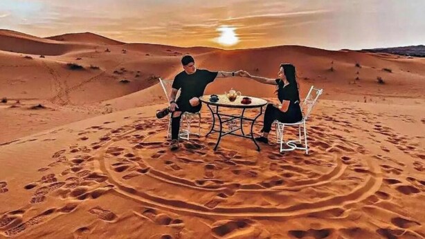 Twee personen zitten aan een tafel in een woestijn, met zandduinen op de achtergrond en een zonsondergang.
