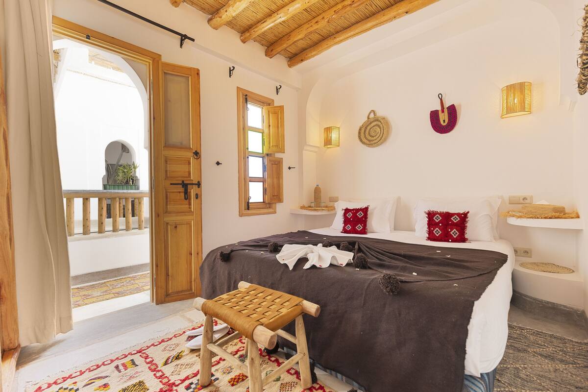 A bedroom with a bed, a wooden bench, a balcony, and a view of the outside.
