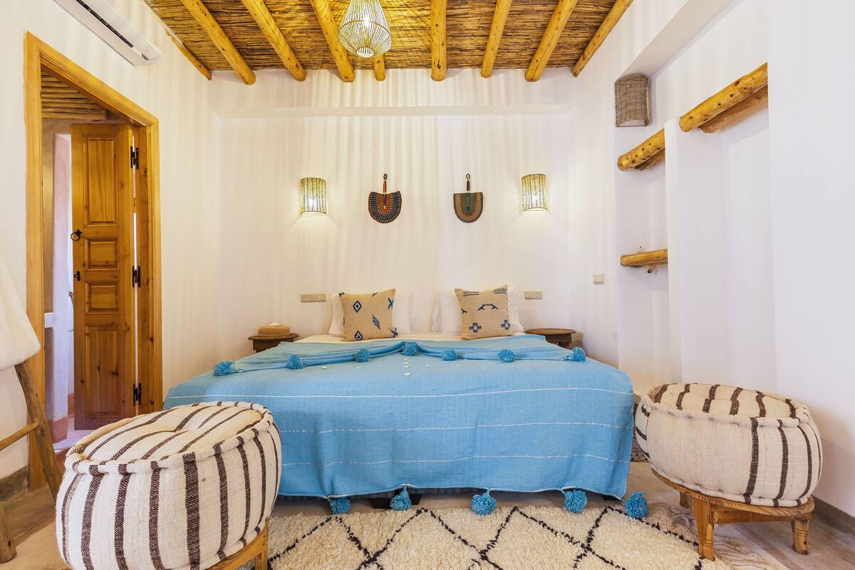 A bedroom with a wooden ceiling, a bed with a blue bedspread, two pillows, a striped ottoman, and a small stool.