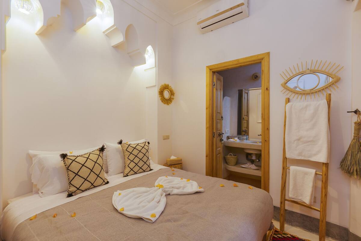 A hotel room with a bed, a wooden ladder with towels, a mirror, and a small table with a basket.
