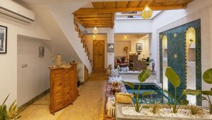 A spacious hallway with a wooden staircase, a pool area with plants, and a dining area with a table and chairs.