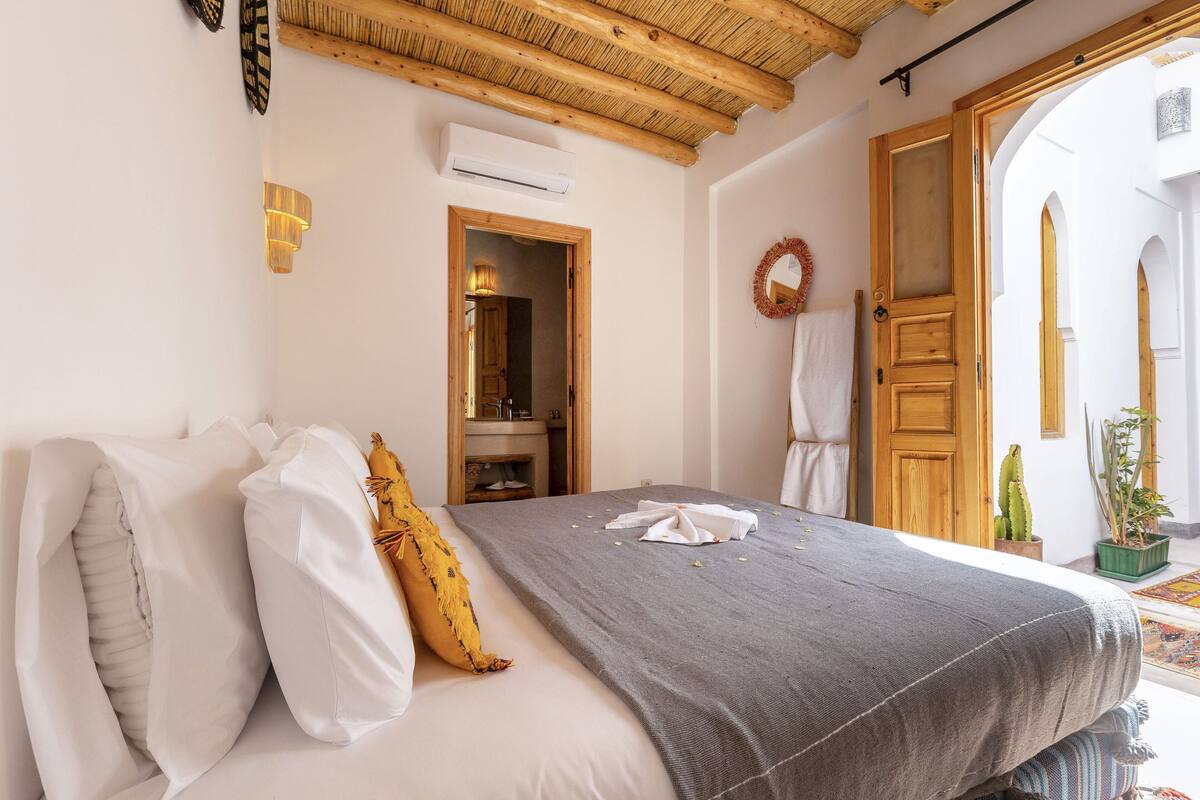 A bedroom with a bed, a wooden ceiling, a door, and a small bathroom visible through an open door.