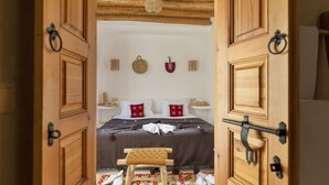 A wooden door leading to a bedroom with a bed, pillows, a small stool, and a wall-mounted shelf.
