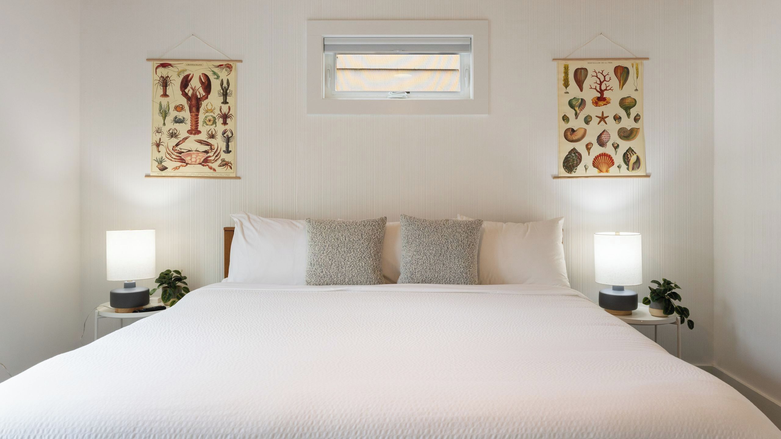 A bedroom with a large bed, two bedside tables with lamps, a window, and two wall-mounted artwork pieces.