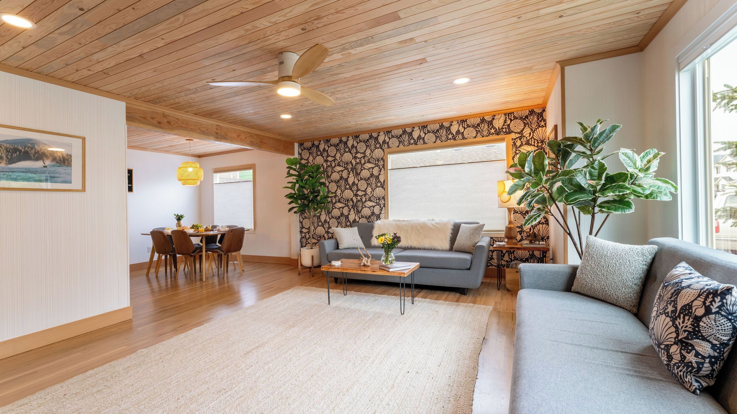 Un soggiorno moderno con soffitto in legno, un divano grigio, un'area pranzo con tavolo e sedie, e un'ampia finestra con persiane.