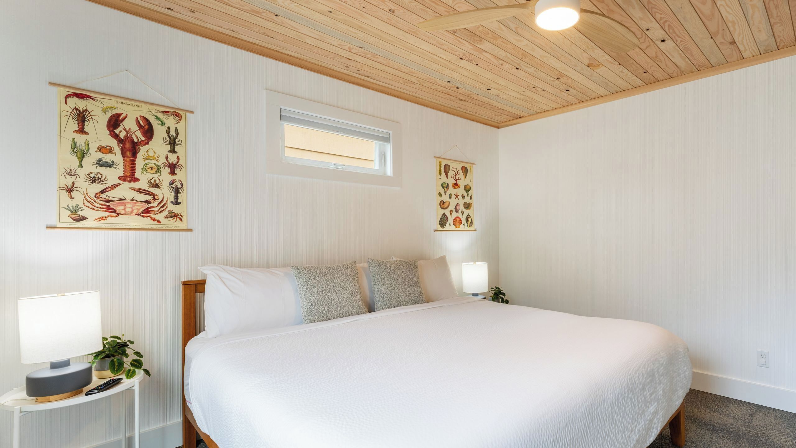 Ein Schlafzimmer mit einem großen Bett, zwei Wandleuchten, einem kleinen Nachttisch mit Lampe sowie zwei gerahmten Pflanzenbildern an der Wand.