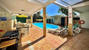 Ein überdachter Außenbereich mit Pool, Liegestühlen, Grill und Esstisch.