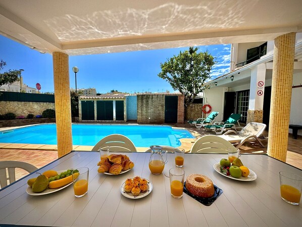 Desayuno junto a la piscina, con una mesa, sillas y vistas a la piscina y edificios al fondo.