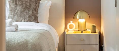 A neatly made bed with a white headboard, a bedside lamp, and a small nightstand with a lamp and a plant.