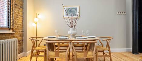A dining area with a round wooden table set for four, surrounded by four matching chairs. A white vase with branches is placed on the table.